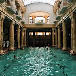 An elegant indoor swimming pool with ornate columns and a high arched glass ceiling. Several people are swimming in the pool, and a few are standing by the edge dressed in white robes. The architectural style is classical with intricate sculptures on the columns. Natural light streams through the skylight, creating reflections on the water.