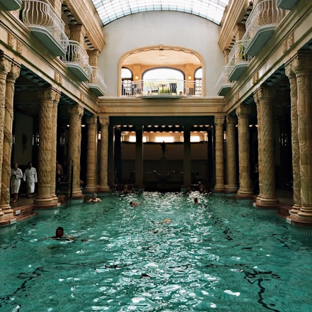 An elegant indoor swimming pool with ornate columns and a high arched glass ceiling. Several people are swimming in the pool, and a few are standing by the edge dressed in white robes. The architectural style is classical with intricate sculptures on the columns. Natural light streams through the skylight, creating reflections on the water.