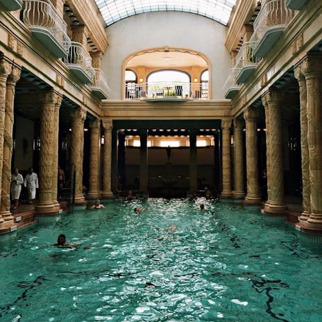 An elegant indoor swimming pool with ornate columns and a high arched glass ceiling. Several people are swimming in the pool, and a few are standing by the edge dressed in white robes. The architectural style is classical with intricate sculptures on the columns. Natural light streams through the skylight, creating reflections on the water.
