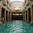 An elegant indoor swimming pool with ornate columns and a high arched glass ceiling. Several people are swimming in the pool, and a few are standing by the edge dressed in white robes. The architectural style is classical with intricate sculptures on the columns. Natural light streams through the skylight, creating reflections on the water.