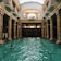 An elegant indoor swimming pool with ornate columns and a high arched glass ceiling. Several people are swimming in the pool, and a few are standing by the edge dressed in white robes. The architectural style is classical with intricate sculptures on the columns. Natural light streams through the skylight, creating reflections on the water.
