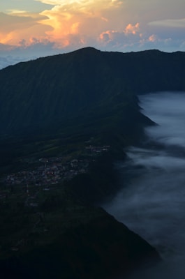 A scenic view of a mountainous landscape with a small village nestled in a valley. Clouds and mist roll through the valley, creating a mystical ambiance. The horizon features a brightly colored sky with orange and pink hues of a sunset.