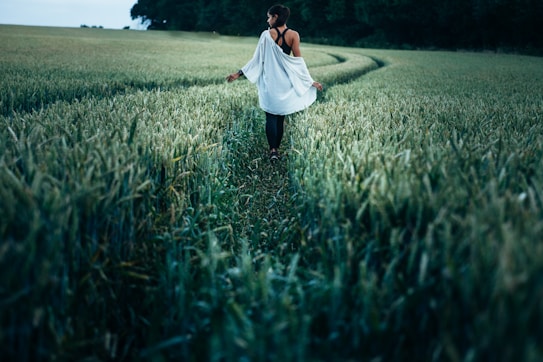 A person walks through a lush, green wheat field, creating a natural path. They are wearing a light, flowing garment and are surrounded by tall crops that sway gently. The atmosphere is calm and serene, with the field extending toward dense, dark trees in the background.