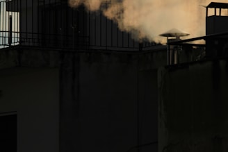 A rooftop scene featuring a chimney emitting smoke against a dark background. The smoke appears light and wispy, contrasting with the shadowed walls of the building. Metal railings and a small illuminated window add architectural elements.
