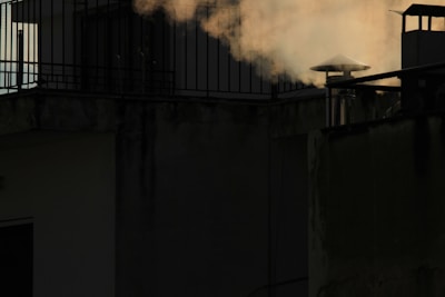 A rooftop scene featuring a chimney emitting smoke against a dark background. The smoke appears light and wispy, contrasting with the shadowed walls of the building. Metal railings and a small illuminated window add architectural elements.