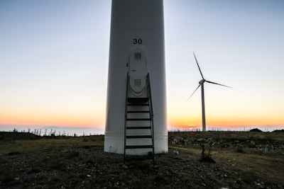 A large wind turbine set against a backdrop of a colorful sunset, with the sky transitioning from soft pastel hues to deeper tones. The ground is rocky and uneven, with sparse vegetation. One prominent turbine is in the foreground with the number '30' on it, and another turbine is visible in the background.