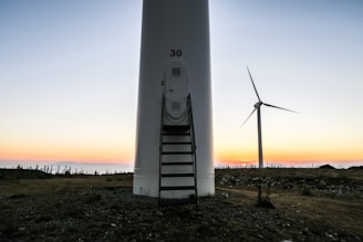 A large wind turbine set against a backdrop of a colorful sunset, with the sky transitioning from soft pastel hues to deeper tones. The ground is rocky and uneven, with sparse vegetation. One prominent turbine is in the foreground with the number '30' on it, and another turbine is visible in the background.