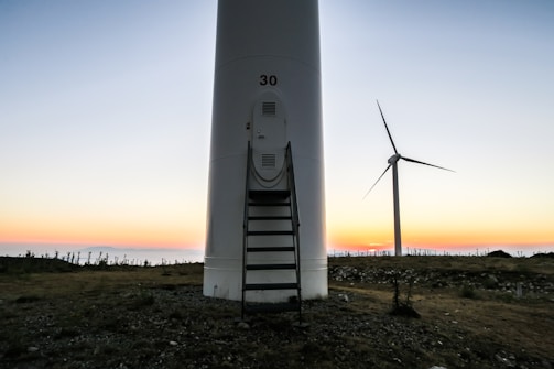 A large wind turbine set against a backdrop of a colorful sunset, with the sky transitioning from soft pastel hues to deeper tones. The ground is rocky and uneven, with sparse vegetation. One prominent turbine is in the foreground with the number '30' on it, and another turbine is visible in the background.
