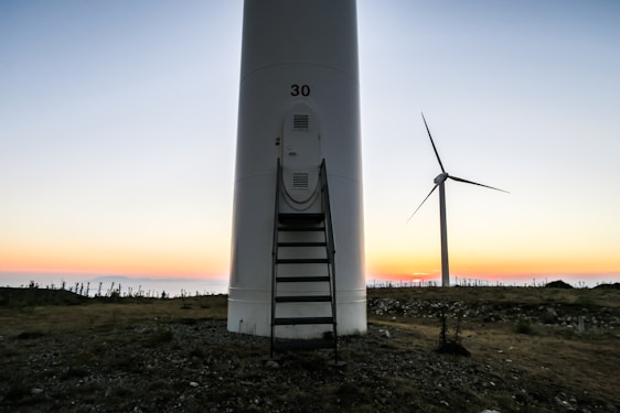 A large wind turbine set against a backdrop of a colorful sunset, with the sky transitioning from soft pastel hues to deeper tones. The ground is rocky and uneven, with sparse vegetation. One prominent turbine is in the foreground with the number '30' on it, and another turbine is visible in the background.