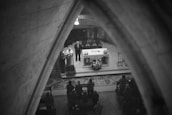 A black and white image captures a religious service inside a church. The scene is viewed through an arched stone structure, focusing on the altar where two clergy members stand. The altar is adorned with candles and a floral arrangement, and is surrounded by a congregation seated on pews.