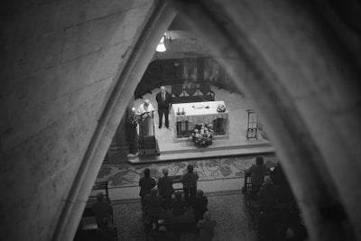 A black and white image captures a religious service inside a church. The scene is viewed through an arched stone structure, focusing on the altar where two clergy members stand. The altar is adorned with candles and a floral arrangement, and is surrounded by a congregation seated on pews.