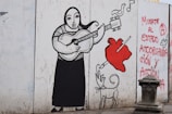 A mural depicts a stylized figure playing a guitar, with a heart being pierced by a pencil nearby. A whimsical dog with exaggerated features is also present. The mural includes birds and musical notes, with graffiti on the side reading, 'Muerte al estado, autoorganización y acción.' The artwork is outlined in bold black lines, with splashes of red.