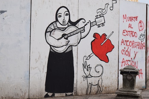 A mural depicts a stylized figure playing a guitar, with a heart being pierced by a pencil nearby. A whimsical dog with exaggerated features is also present. The mural includes birds and musical notes, with graffiti on the side reading, 'Muerte al estado, autoorganización y acción.' The artwork is outlined in bold black lines, with splashes of red.