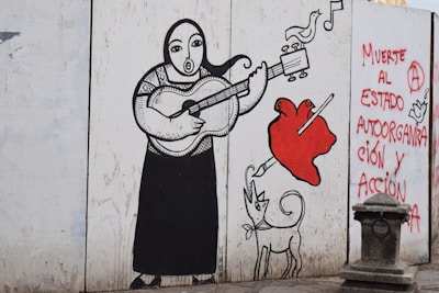 A mural depicts a stylized figure playing a guitar, with a heart being pierced by a pencil nearby. A whimsical dog with exaggerated features is also present. The mural includes birds and musical notes, with graffiti on the side reading, 'Muerte al estado, autoorganización y acción.' The artwork is outlined in bold black lines, with splashes of red.