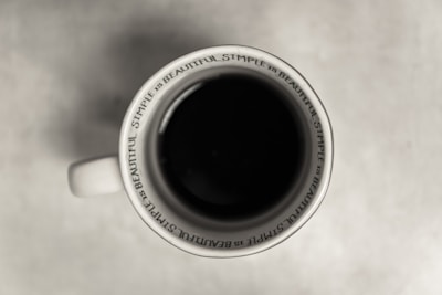 A top-down view of a white mug filled with a dark liquid, likely coffee. The mug's interior rim has the phrase 'Simple is Beautiful' repeated multiple times. The background is a neutral, light surface.