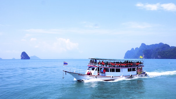 A large tour boat filled with passengers wearing orange life jackets sails on a vast expanse of calm blue ocean. The boat is white with a blue roof and adorned with flags. In the background, several small rocky islands are scattered across the horizon under a bright blue sky with a few clouds.