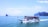 A large tour boat filled with passengers wearing orange life jackets sails on a vast expanse of calm blue ocean. The boat is white with a blue roof and adorned with flags. In the background, several small rocky islands are scattered across the horizon under a bright blue sky with a few clouds.