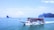 A large tour boat filled with passengers wearing orange life jackets sails on a vast expanse of calm blue ocean. The boat is white with a blue roof and adorned with flags. In the background, several small rocky islands are scattered across the horizon under a bright blue sky with a few clouds.