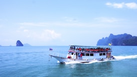 A large tour boat filled with passengers wearing orange life jackets sails on a vast expanse of calm blue ocean. The boat is white with a blue roof and adorned with flags. In the background, several small rocky islands are scattered across the horizon under a bright blue sky with a few clouds.