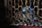 A small rodent, likely a rat, is inside a cage with a metal mesh design. The lighting is dim, creating shadows, and the metal has a rusted appearance. Blue markings are visible on the rat's fur, adding a unique element to the scene.
