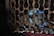 A small rodent, likely a rat, is inside a cage with a metal mesh design. The lighting is dim, creating shadows, and the metal has a rusted appearance. Blue markings are visible on the rat's fur, adding a unique element to the scene.