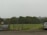 A rural scene with overcast skies and a green field bordered by dense trees. In the foreground, there's a car parked on a dirt lot next to a large garbage bin. A small pile of debris and some scattered items are visible nearby.