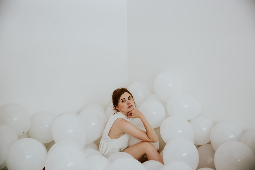 白いノースリーブのトップスを着た女性が、白い風船に囲まれた床に座り、白い塗りつぶされた部屋の中で