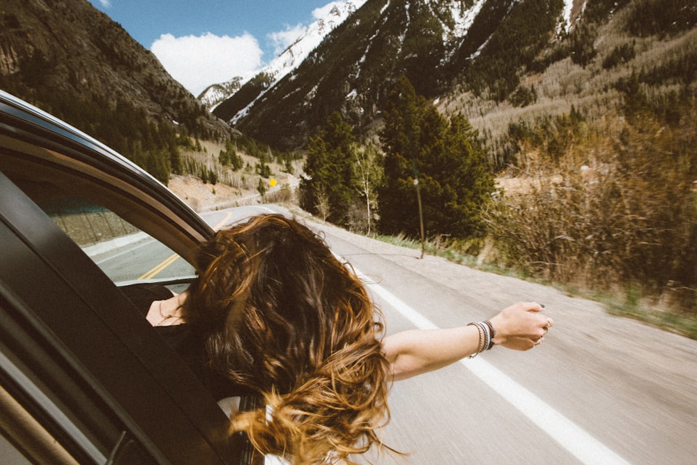 Mujer que viaja en un vehículo sacando la cabeza y el brazo derecho por la ventanilla mientras viaja por la carretera