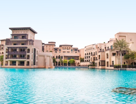 A luxurious architectural complex with beige buildings featuring arches and balconies sits beside a large, tranquil pool of clear blue water. Palm trees and well-manicured greenery accent the area, under a clear blue sky.