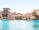 A luxurious architectural complex with beige buildings featuring arches and balconies sits beside a large, tranquil pool of clear blue water. Palm trees and well-manicured greenery accent the area, under a clear blue sky.