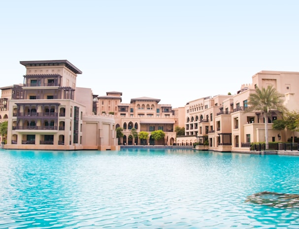 A luxurious architectural complex with beige buildings featuring arches and balconies sits beside a large, tranquil pool of clear blue water. Palm trees and well-manicured greenery accent the area, under a clear blue sky.