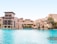 A luxurious architectural complex with beige buildings featuring arches and balconies sits beside a large, tranquil pool of clear blue water. Palm trees and well-manicured greenery accent the area, under a clear blue sky.