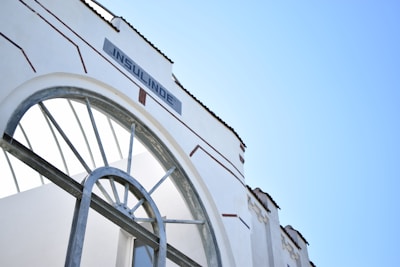 An architectural detail of a white building with an arched window and metallic framing. The word 'INSULINDE' is displayed prominently on a sign above the arch. The sky is clear and blue, adding to the clean and modern aesthetic.