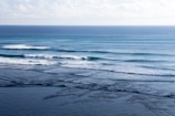 Waves gently roll towards the shore, creating a rhythmic pattern across the ocean surface. Several surfers can be seen waiting to catch the next wave, adding a sense of life and activity to the scene. The horizon is visible in the distance under a sky dotted with soft clouds.