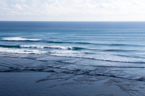 Waves gently roll towards the shore, creating a rhythmic pattern across the ocean surface. Several surfers can be seen waiting to catch the next wave, adding a sense of life and activity to the scene. The horizon is visible in the distance under a sky dotted with soft clouds.