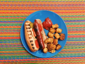 A grilled hot dog in a bun with mustard, pickle, and tomato slices is placed on a blue plate alongside a portion of tater tots and a dollop of ketchup. The background consists of a colorful woven mat with vibrant stripes.