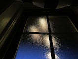 A glass window is observed with frosted texture, dimly lit from behind. The light creates a warm glow, contrasting with the dark framework of the window. Outside, a deep blue hue suggests twilight or early morning.