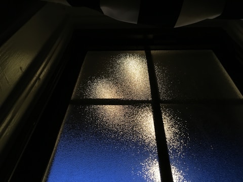 A glass window is observed with frosted texture, dimly lit from behind. The light creates a warm glow, contrasting with the dark framework of the window. Outside, a deep blue hue suggests twilight or early morning.