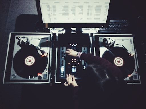 A person is adjusting controls on a DJ mixer, with two turntables on either side and a monitor displaying waveforms or music tracks above. The scene is dimly lit, creating a focused, intense atmosphere.
