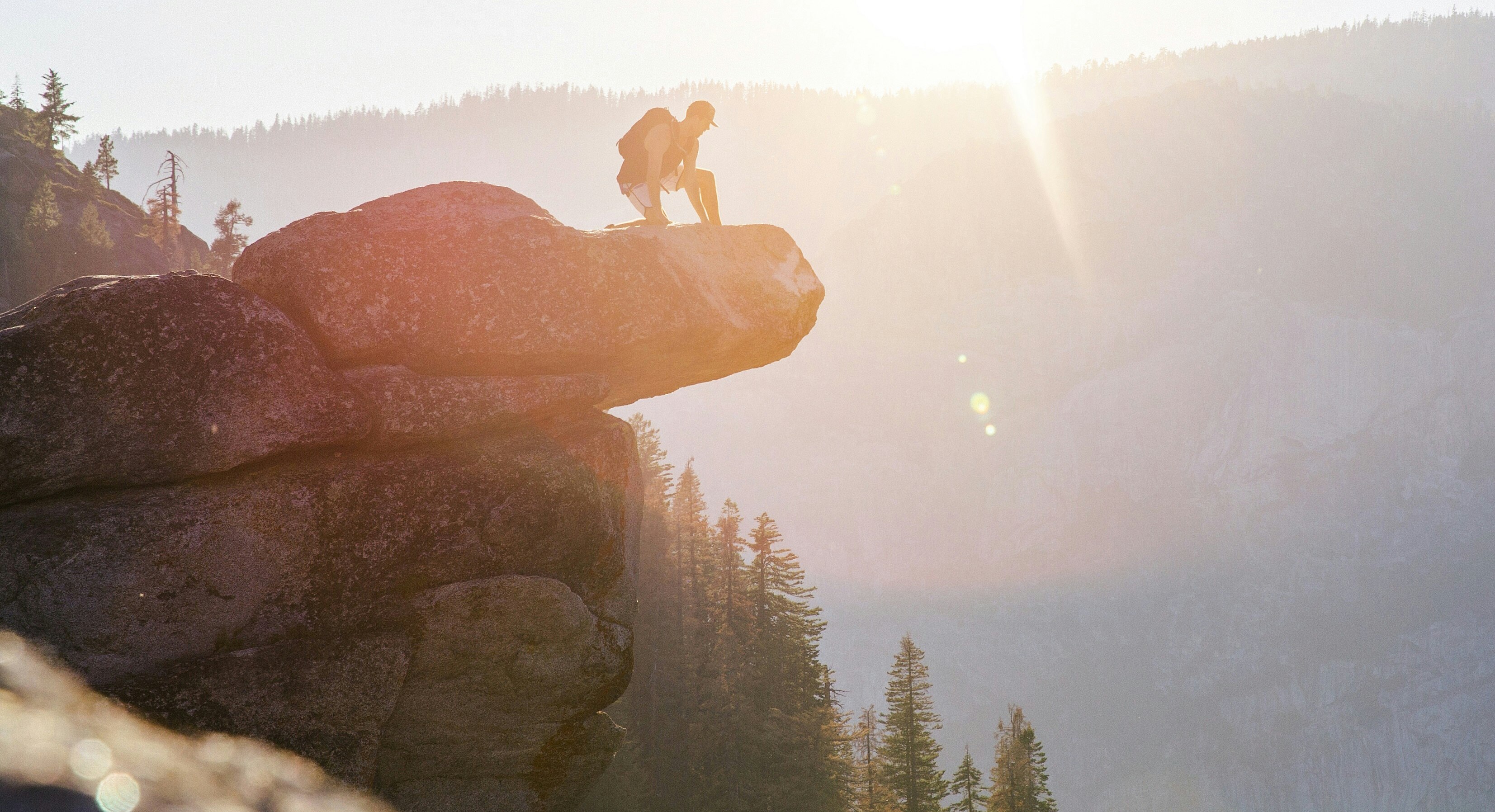 A person kneeling on the ledge of a rock with the sunset in the background at Glacier Point