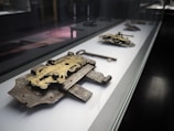 A display case holds a collection of intricate, antique locks and a key, reflecting historical craftsmanship with metalwork in a museum-like setting. The locks are ornate, featuring detailed engravings and a patina finish, with the key positioned prominently next to a lock.