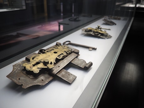 A display case holds a collection of intricate, antique locks and a key, reflecting historical craftsmanship with metalwork in a museum-like setting. The locks are ornate, featuring detailed engravings and a patina finish, with the key positioned prominently next to a lock.