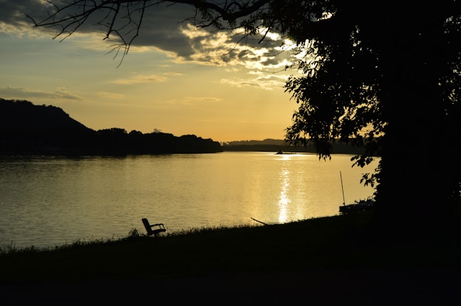 A serene lakeside scene during sunset, with golden sunlight reflecting off the water. A silhouetted tree provides a natural frame, and an empty bench faces the tranquil view. The sky is partially cloudy, casting soft shadows on the distant hills.