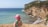 A child wearing a pink hat and a floral shirt is standing near a coastal cliff, overlooking a turquoise sea. The rugged coastline features a sandy beach with gentle waves lapping against the shore. Dense greenery covers the cliffside, and the clear sky above suggests a sunny day.