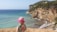 A child wearing a pink hat and a floral shirt is standing near a coastal cliff, overlooking a turquoise sea. The rugged coastline features a sandy beach with gentle waves lapping against the shore. Dense greenery covers the cliffside, and the clear sky above suggests a sunny day.