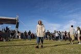 A young person wearing a hoodie and shorts is walking on a grassy field in front of a crowd gathered around a stage setup. The event appears to be outdoors, with a blue sky overhead. The stage has speakers and equipment visible on it, with several people nearby. In the distance, more people are gathered, some standing and some sitting on the grass.