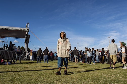 A young person wearing a hoodie and shorts is walking on a grassy field in front of a crowd gathered around a stage setup. The event appears to be outdoors, with a blue sky overhead. The stage has speakers and equipment visible on it, with several people nearby. In the distance, more people are gathered, some standing and some sitting on the grass.