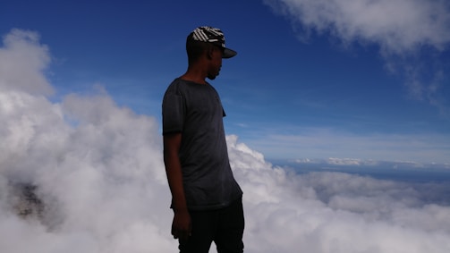 A person stands near the edge of a cloud-covered landscape, looking out at the vast expanse of clouds and the blue sky above. The individual is wearing a dark gray T-shirt and a black hat with a white pattern.