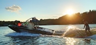 A person is wakeboarding behind a motorboat on a body of water during sunset. The motorboat is creating waves, and there are trees and a partly cloudy sky in the background. The sun is low on the horizon, casting a warm glow over the scene.
