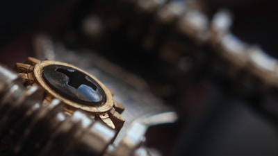 A close-up view of a metallic object featuring a gold and black design. The object includes a gear-like structure with a glossy, black surface in the center, surrounded by a ring of gold. The background is blurred, focusing attention on the intricate details of the metal and the reflective nature of the black surface.